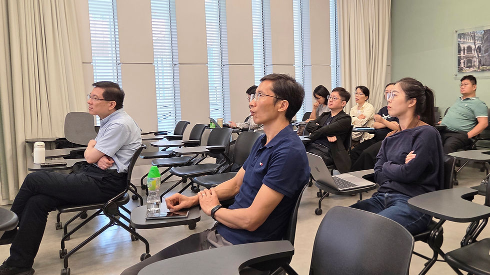 Adults sit in a bright classroom, listening to a presentation; laptops and water bottles on desks, focused expressions.