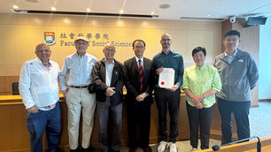 Seven people posing in a university lecture hall before a Faculty of Social Sciences sign; one man holds a certificate.