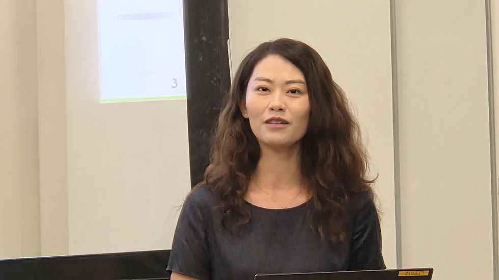 Woman with wavy dark hair stands in a meeting room before a projected slide showing 3, looking attentive and neutral.