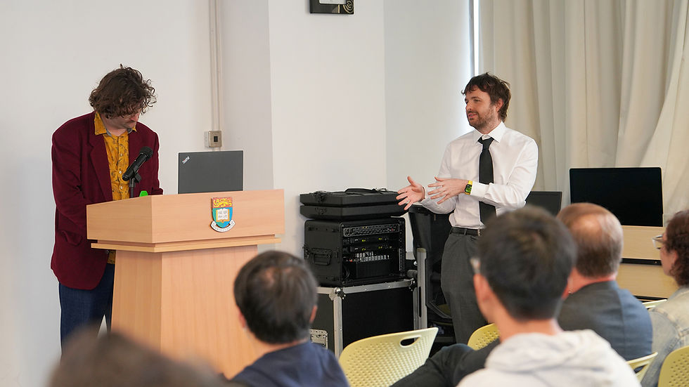 Two men present in a classroom, one at a podium and one gesturing, while an audience listens to a talk.
