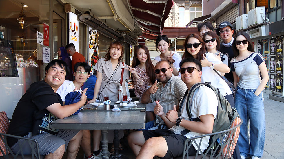 Smiling group of friends posing and giving thumbs up at an outdoor cafe on a city street, with awnings and posters behind them.