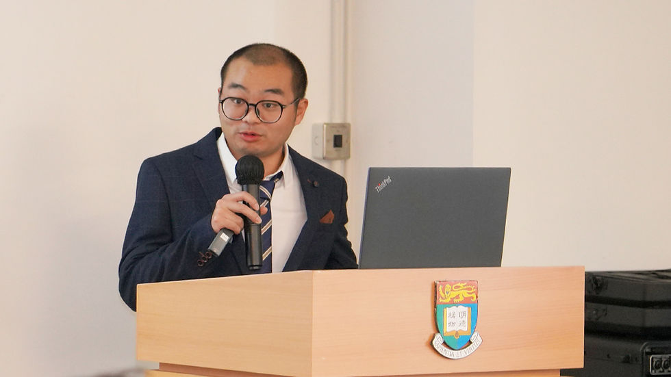 Man in a suit speaks at a podium with a microphone and laptop in a plain room, looking formal and focused.