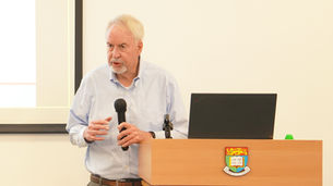 Older man speaks at a podium with a microphone and laptop, in front of a projection screen and university crest.