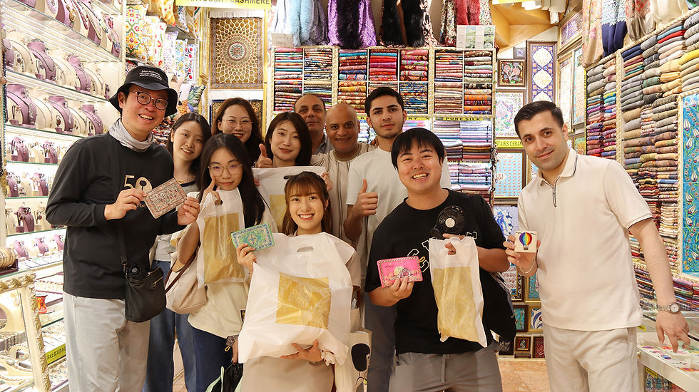 Smiling group of shoppers poses in a colorful textile shop, holding bags and small souvenirs amid stacked folded fabrics.