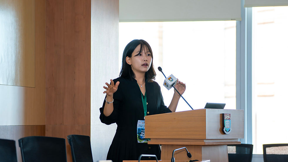 Woman speaks at a podium in a bright conference room, gesturing beside a microphone, with a university crest on the lectern.