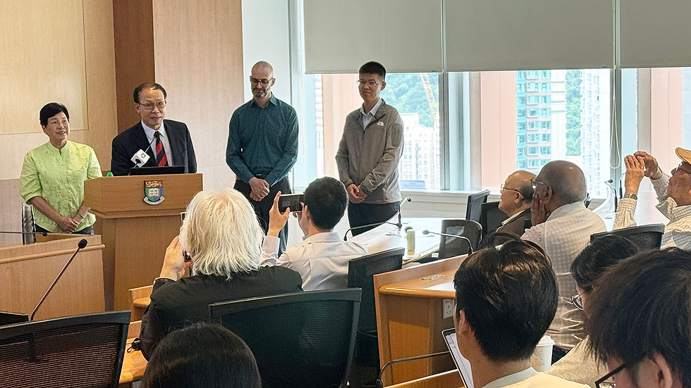 People at a formal meeting in a bright conference room, speaker at podium while audience takes photos, with city view outside.