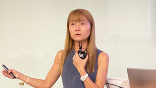 Woman giving a presentation, holding a microphone and remote beside a Dell laptop, against a plain white wall, focused expression