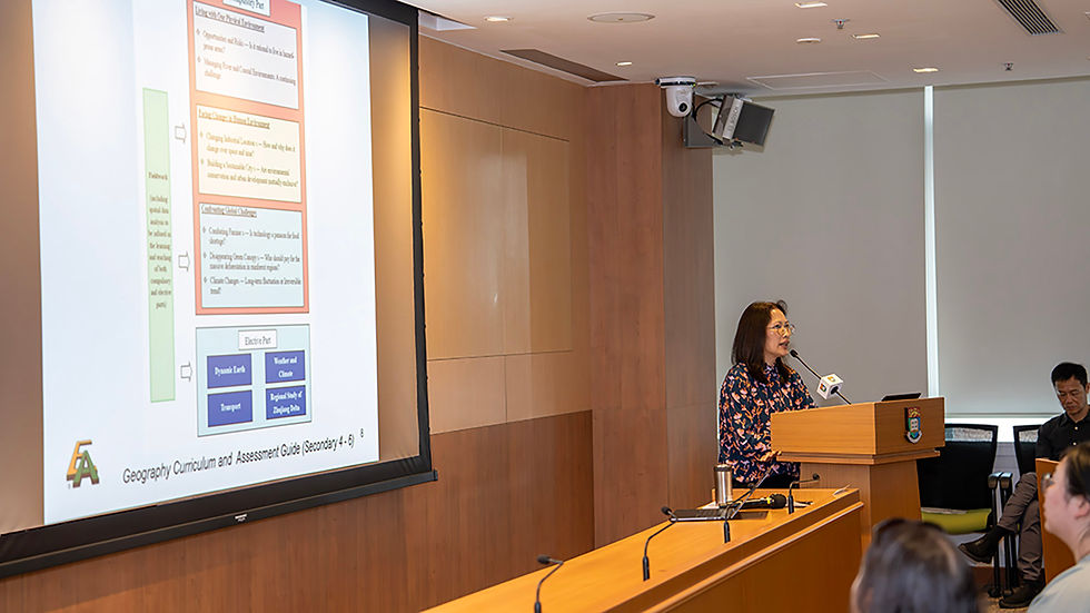Woman speaks at podium during a presentation in a conference room; slide shows Geography Curriculum and Assessment Guide on screen.