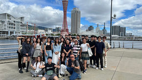 Group of young adults posing by a waterfront railing, making peace signs, with a red tower and city skyline under cloudy skies.