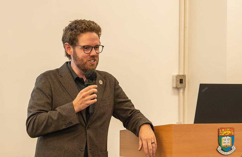 Man with glasses speaks into a microphone at a podium beside a ThinkPad laptop, with a crest reading Sapientia et Virtus.