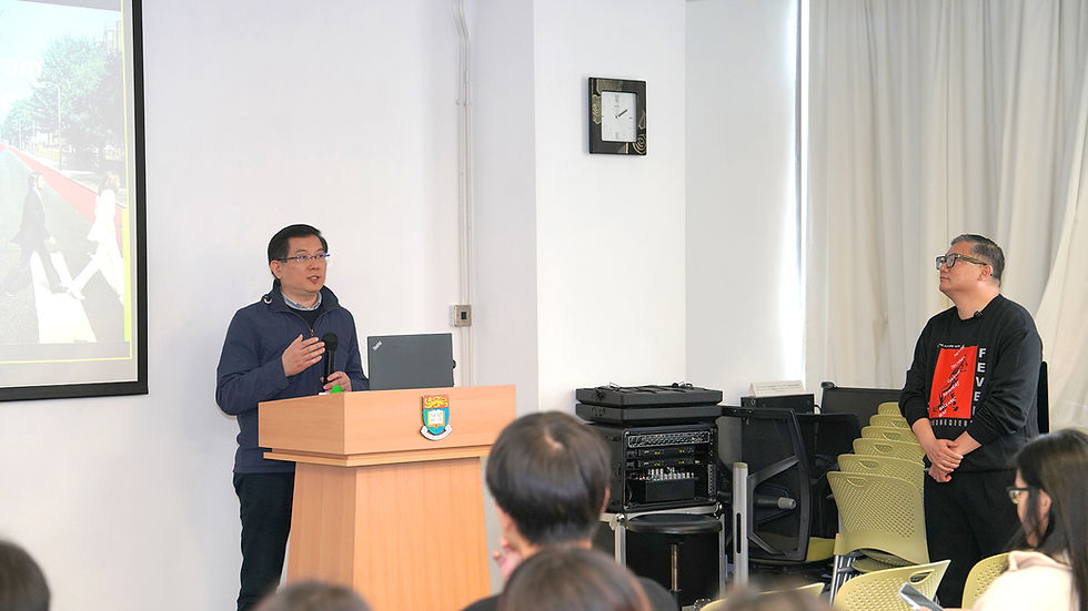 Speaker at a podium giving a talk to seated attendees in a bright classroom, with a projected slide and clock on the wall.