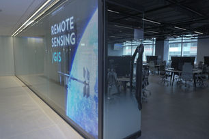 Glass-walled office with large screen reading REMOTE SENSING GIS, empty desks and monitors in a modern workspace.