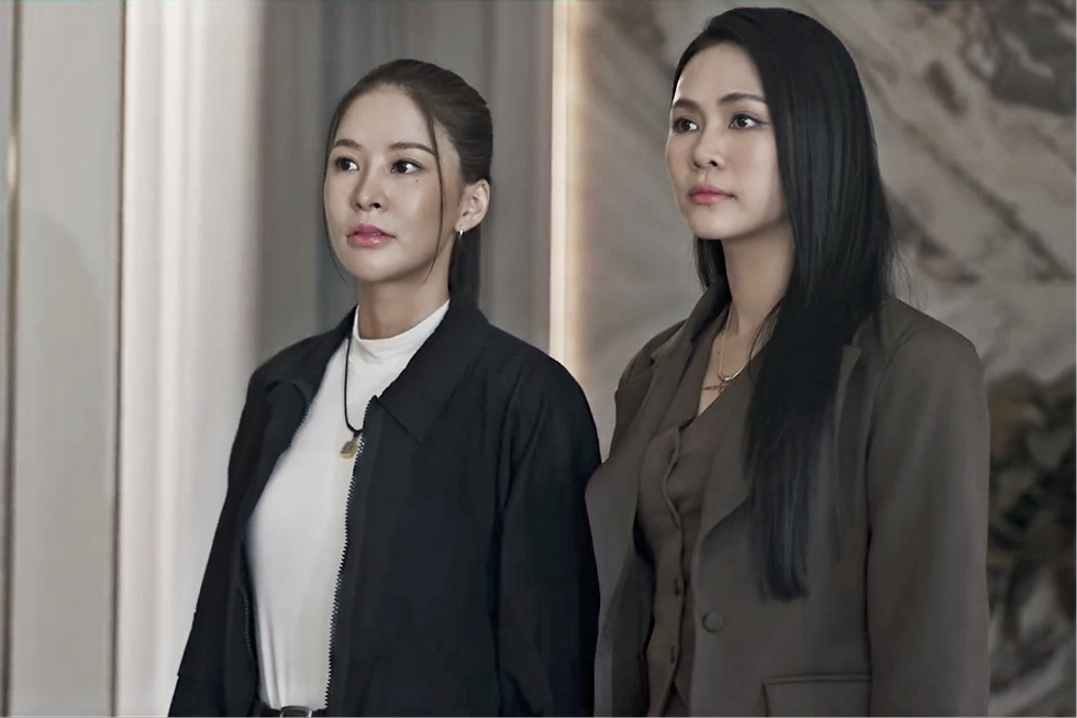Peem, wearing a black jacket, stands beside Bulan in a tailored brown suit as they prepare to meet Peem’s mother at her family home in Khom Khlang Episode 8.