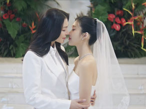 Arisa and Lalin share an intimate moment at their wedding ceremony, leaning in for a kiss after exchanging vows, surrounded by lush floral arrangements.