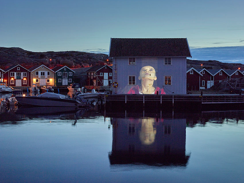 Dusk over a harbor village with boats and colorful houses, a large face projected on a white building, reflected in calm water.
