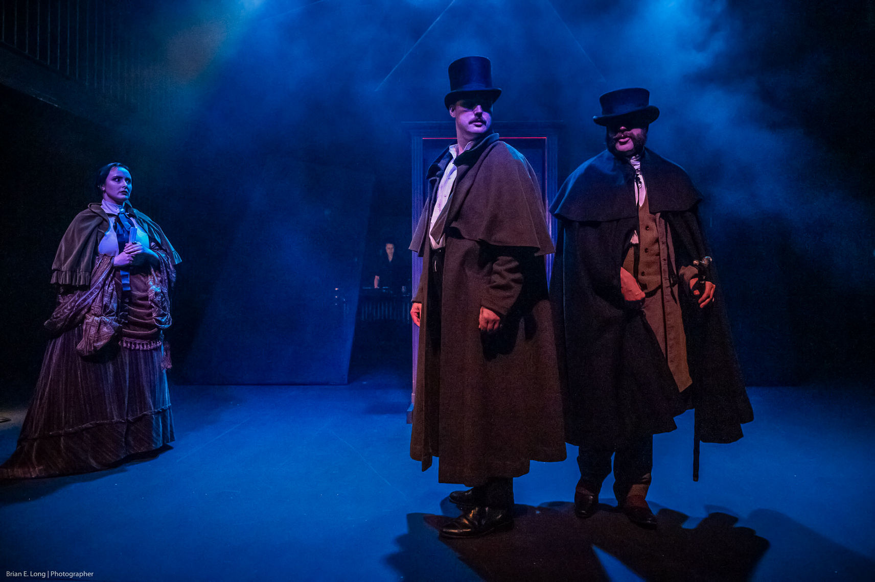 Two men in trench coats and hats stand at the forefront. Behind them, the air is foggy and blue. A woman stands to the side look at them concerned. 