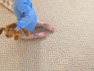 Baby in a blue onesie sits on a woven carpet flooring, holding a brown stuffed dog toy.