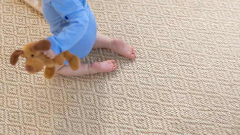 Baby in a blue onesie sits on a woven carpet flooring, holding a brown stuffed dog toy.