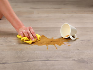 Hand cleaning coffee spill on wooden floor with yellow cloth. An overturned white mug lies nearby. Bright, clean, domestic setting.