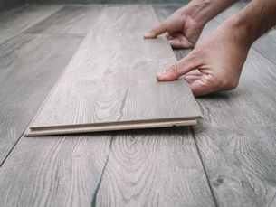 Hands are installing a light gray wood LVT flooring piece in a room. The floor has a natural wood grain pattern.