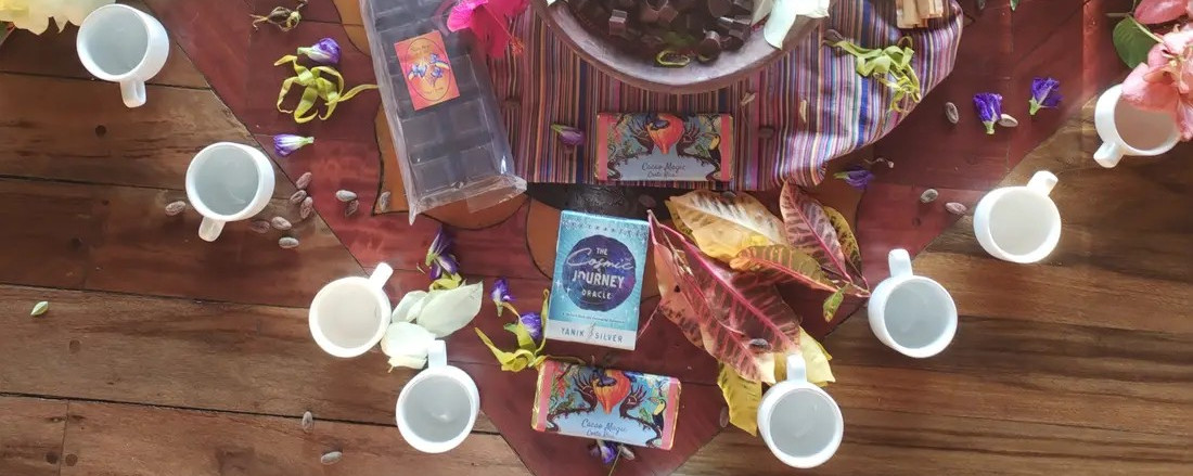 An overhead view of a cacao ceremony: a ring of white cups around a heart-shaped bowl of cacao, a pod, flowers and leaves on a wooden floor
