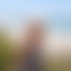 An older woman smiling at the camera, in black and white swimsuit, with blue sea and blue sky and white sand and beach greenery around her.