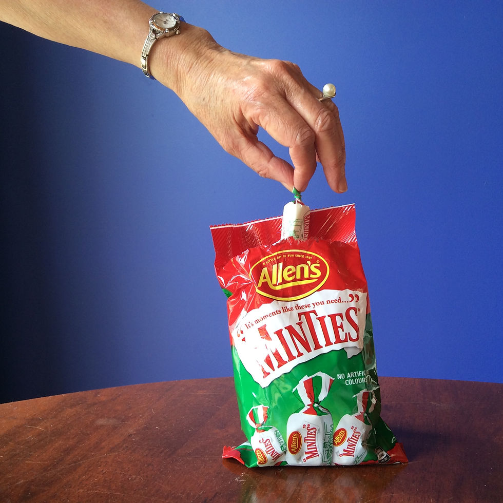 A hand reaching into a packet of Allen's Minties and removing one. Blue wall, wooden table. Pearl ring on her finger, silver watch on her hand.