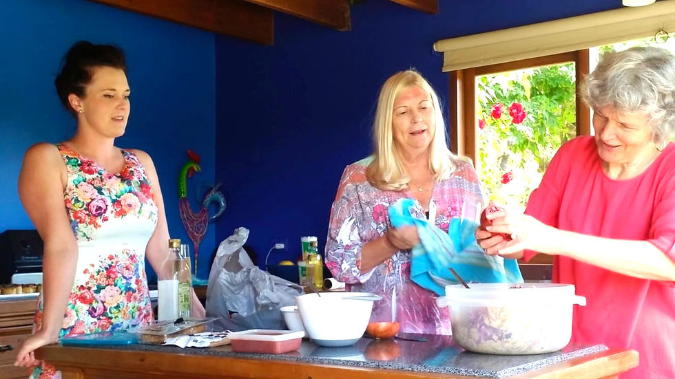 Young woman in sleeveless, flowery dress, and two older women gather around a kitchen counter, making a salad.