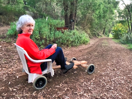 An older woman in a red top and dark blue jeans on a home-made hill trolley at the top of a driveway lined with trees. She's about to take off.