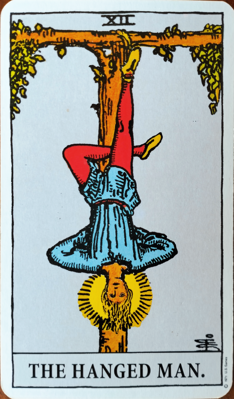 The Hanged Man of the Rider-Waite Tarot, which is a man in red tights and a blue tunic suspended upside down on branches forming a T-shape.