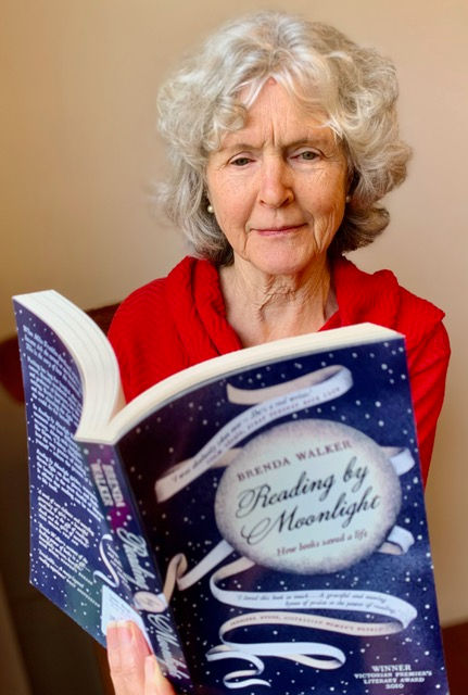 An older woman with grey hair and red jumper, reading a book called "Reading by Moonlight", by Brenda Walker.