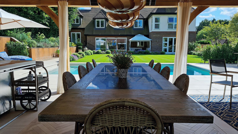Pool outbuilding with wood table with glass inlay seating 10 people. There is a bar to the left and the pool is outside in the sunshine.