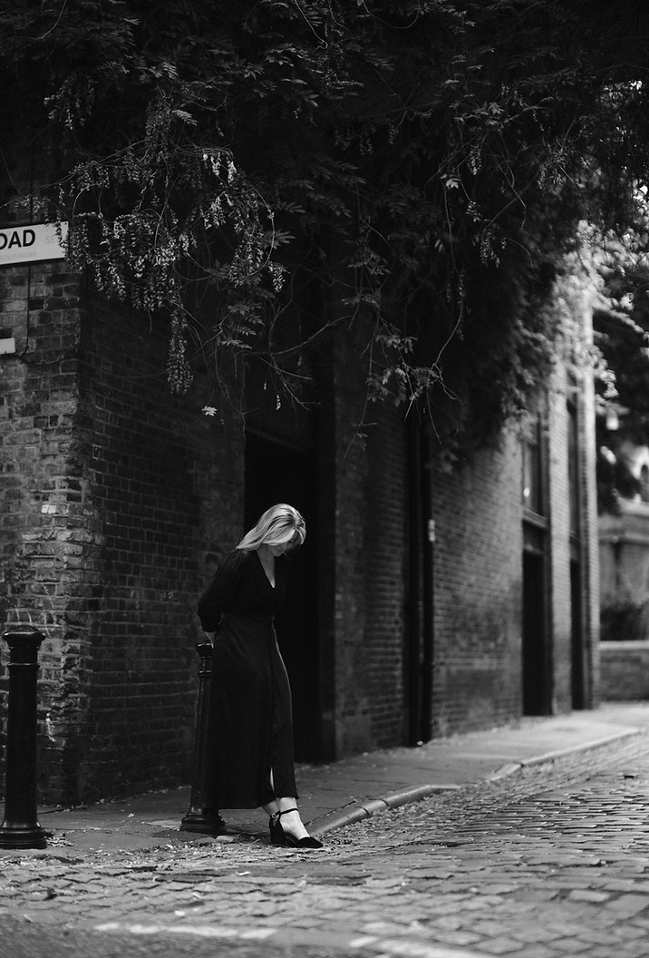 Ella is pictured in black and white wearing a satin dress and velvet heels, leaning against a post on a cobbled lane