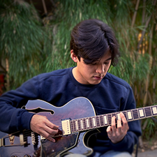 Headshot of Chris Lee, wearing a navy blue sweatshirt and playing the guitar.