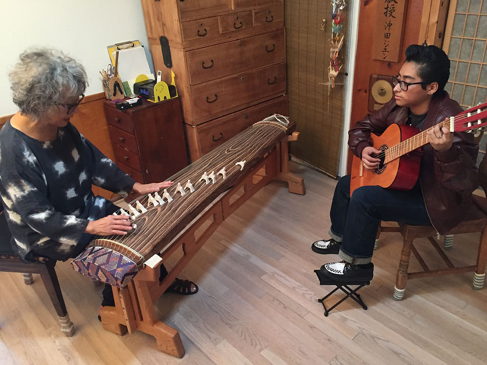 June Kuramoto, wearing a black sweater with white circles players her koto with Jayleen Montoya, wearing a brown leather jacket and black pants, plays her guitar.