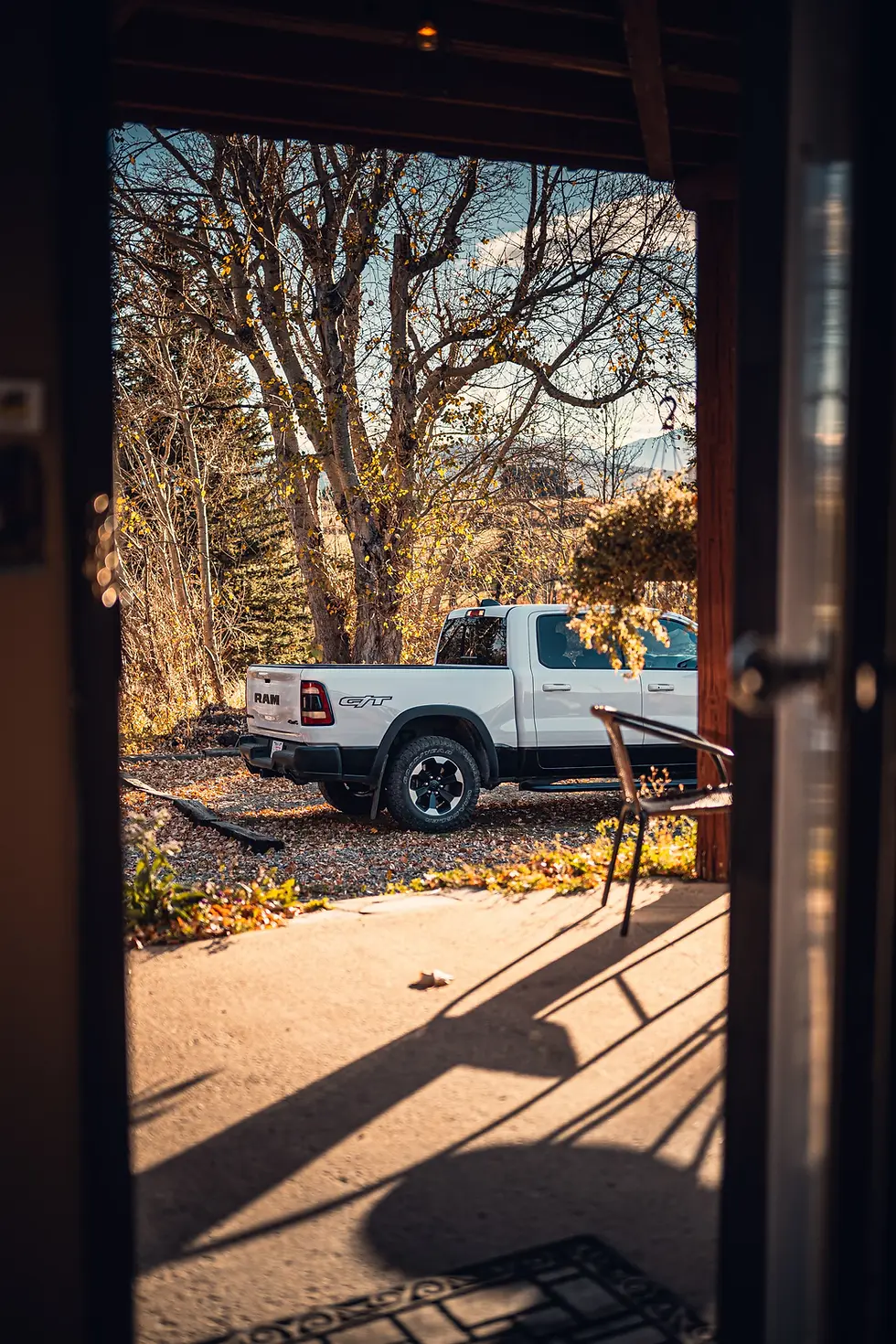 Witte pick-uptruck geparkeerd in een herfstige tuin met kale bomen. Zonnige dag, zicht vanuit een deur. Schaduw en blauwe lucht zichtbaar.