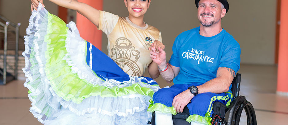 Homem cadeirante com roupa de festa junina com a logo "street cadeirante" e uma bailarina de saia rodada ao seu lado. ambos sorriem e olham para a câmera