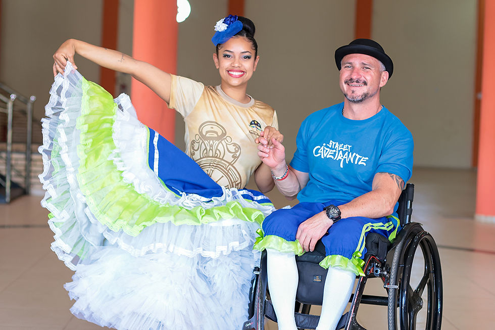 Homem cadeirante com roupa de festa junina com a logo "street cadeirante" e uma bailarina de saia rodada ao seu lado. ambos sorriem e olham para a câmera