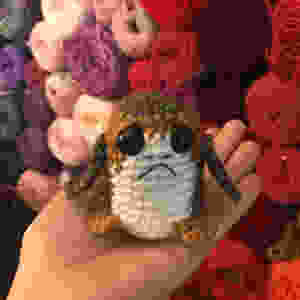 A hand is holding a small crocheted Star Wars Porg in front of a wall of yarn balls