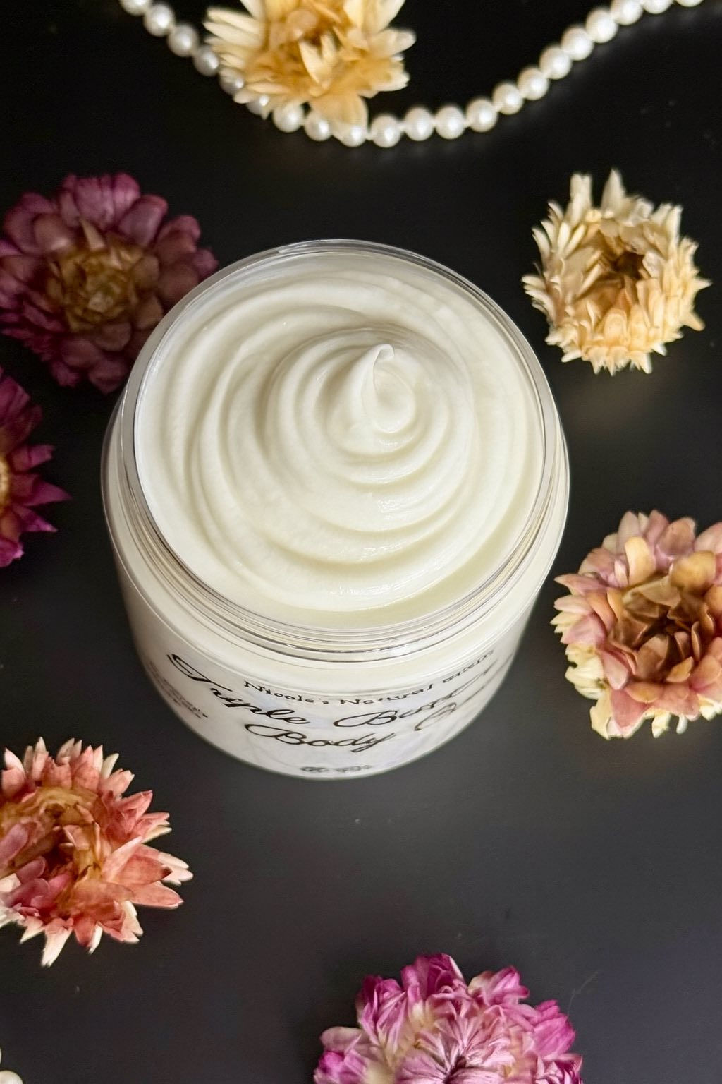 A top-down view of an open white cosmetic jar filled with a thick, white cream featuring a peaked swirl pattern, surrounded by four dried flowers in shades of orange and pink on a dark background.