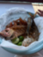 Fried fish and prawns in a blue basket lined with paper, garnished with limes, on a table. A hand is in the background.