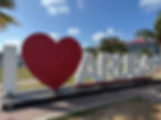 Large "I ❤️ ARUBA" sign with a red heart on a sunny day. Cruise ship in the background, surrounded by palm trees and blue sky.