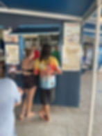 Two people stand at a gelato shop counter under a blue awning. Menus and signs are visible. The setting is casual and relaxed.