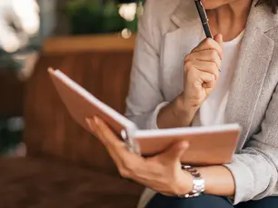A woman in a gray blazer sits on a brown sofa, reading a notebook and holding a pen thoughtfully, in a cozy indoor setting.