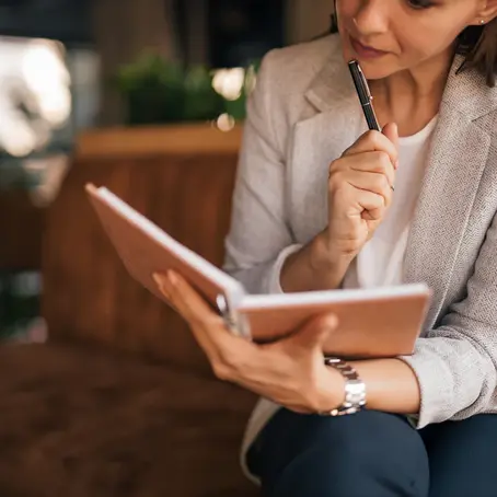 A woman in a gray blazer sits on a brown sofa, reading a notebook and holding a pen thoughtfully, in a cozy indoor setting.