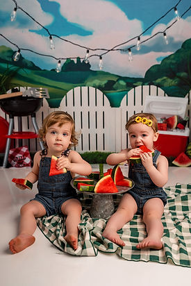 Boy and girl twins sit eating watermelon in front of a BBQ backdrop.