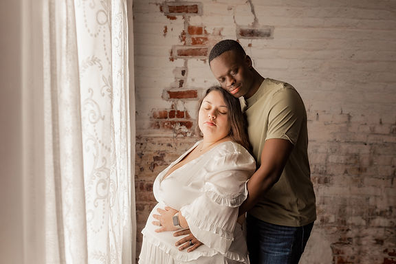 Couple leans together with hands on pregnant belly in front of a window.