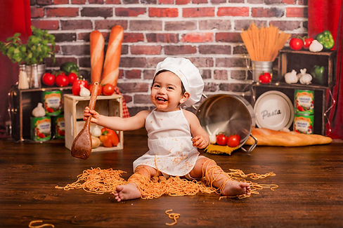 Toddler smiles wearing an apron and chef hat, holding spoon, and covered in spaghetti.