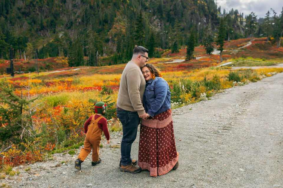 Family surrounded by fall foliage - mom and dad embrace while toddler runs around them. 