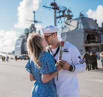 Man in military uniform kissing his wife in front of Naval ship